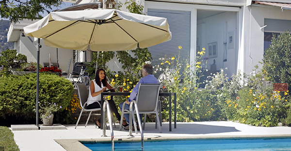 Farah Guignard de Clémo Immobilier conseillant un client sous un parasol au bord d'une piscine, avec un jardin fleuri derrière.